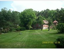 Eine weitläufige Rasenfläche mit Bäumen und Sträuchern, im Hintergrund ein Backsteinhaus mit roten Dachziegeln und Fensterläden sowie ein kleines Nebengebäude mit ähnlichem Dach. Der Bereich ist von dichtem Wald umgeben, und ein einzelnes Schwein steht auf der Wiese.