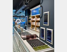 Ein moderner Eissalon mit einer langen Theke aus weißem Holz, darauf Metallbehälter für Eis, gestapelte Gläser und kleine Flaschen. Regale im Hintergrund enthalten Eisbecher und Zubehör, während digitale Preistafeln an der dunkelblauen Wand hängen. Durch ein großes Fenster ist eine Straße mit Fußgängern sichtbar.