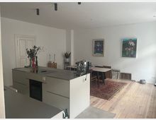 Moderne Küche und Wohnbereich mit heller Holzbodenoptik, einer zentralen Kücheninsel mit eingebautem Herd und Backofen. An der Wand befinden sich zwei bunte Gemälde, und ein Esstisch mit Stühlen steht auf einem roten Teppich.