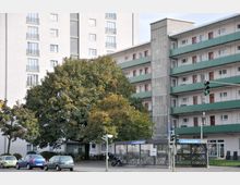 Wohngebäude mit Balkonen und grünen Brüstungen, flankiert von einem großen Laubbaum im Vordergrund. U-Bahn-Eingang „Berliner Straße“ sowie eine Verkehrsampel und geparkte Autos befinden sich im Straßenzug davor.