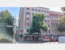 Straßenecke mit einem fünfstöckigen Gebäude in rosafarbenen und cremeweißen Fassadenfarben; im Erdgeschoss befindet sich ein Restaurant mit Schild "Lalibela", daneben weitere Läden. Ein großer Baum steht vor dem Gebäude. Straßenschild "Ohlauer Straße" links im Bild.