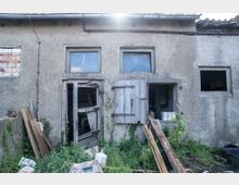 Eine verlassene, heruntergekommene Betonhütte mit beschädigten Holzfensterläden, einer zerbrochenen Tür und überwuchertem Unkraut im Vordergrund. Teile von Baumaterialien und Schutt liegen verstreut um das Gebäude.