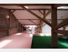 Loftartiger Raum mit freiliegenden Backsteinwänden und sichtbaren Holzbalken. Der Boden ist teils rosafarben, teils mit grünem Teppich bedeckt, und das Licht dringt durch Dachfenster, die schräge weiße Deckenflächen unterbrechen.