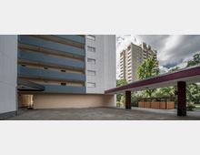 Mehrgeschossige Wohngebäude mit Balkonen und schlichter Fassade, umgeben von Bäumen und einem gepflasterten Vorplatz. Im Vordergrund ist eine überdachte Durchfahrt mit roten Säulen zu sehen.