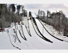 Verschneite Skisprunganlage mit mehreren Schanzen unterschiedlicher Größe, umgeben von winterlich kahlen Bäumen in einer bewaldeten Hügellandschaft. Im Hintergrund sind ein Sprungrichterturm und Flutlichtmasten sichtbar.