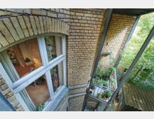 Blick auf eine Ziegelfassade mit einem großen Fenster, das einen Innenraum mit hölzernem Boden und Dekoration zeigt. Rechts daneben ein kleiner, bepflanzter Balkon mit Töpfen und Kräutern, umgeben von Metallstreben und mit Bäumen im Hintergrund.