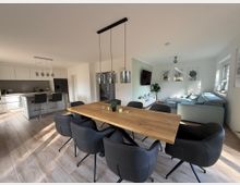 Modernes, offen gestaltetes Wohnzimmer mit Essbereich und Küche. Ein rechteckiger Holztisch mit schwarzen Stühlen steht im Vordergrund, dahinter eine helle Küche mit Theke und Barhockern sowie ein Wohnzimmer mit heller Couch, Wanddekorationen und großen Fenstern, die viel Tageslicht hereinlassen.