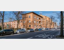 Mehrstöckiges Wohngebäude aus orangefarbenem Putz mit rechteckigen Fenstern, gelegen an einer Straße mit markierter Busspur. Vor dem Gebäude stehen geparkte Autos, und große Bäume säumen die Straße.