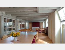 Ein heller Gemeinderaum mit Holzmöbeln und Holzboden, einem großen Tisch mit Stühlen im Vordergrund und einem kleinen Altar mit Vorhängen und Kreuz im Hintergrund. Der Raum verfügt über freiliegende Deckenbalken, seitliche Fenster und eine Vase mit Sonnenblumen auf dem Tisch.