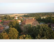 Luftaufnahme eines ländlichen Dorfes mit einem zentralen historischen Fachwerkgebäude, umgeben von Bäumen und kleineren Wohnhäusern. Im Hintergrund sind Felder und ein dichter Wald zu erkennen.