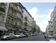 Straßenszene in einer Stadt mit mehrstöckigen Wohn- und Geschäftshäusern aus der Gründerzeit, verziert mit Stuckfassaden und Balkonen. Am Straßenrand sind geparkte Autos, und im Erdgeschoss befinden sich Geschäfte und Cafés; der Himmel ist leicht bewölkt.