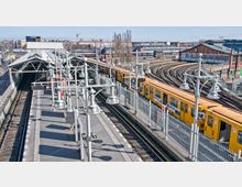 Oberirdische U-Bahn-Station mit einem gelben Zug, der entlang eines Bahnsteigs steht. Im Hintergrund sind mehrere Gleise, ein modernes Bahnhofsgebäude und städtische Gebäude zu sehen.