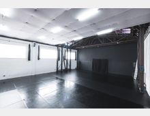 Ein großer heller Raum mit schwarzem Boden, weißen Wänden und mehreren rechteckigen Fenstern, die mit Vorhängen versehen sind. Die Decke ist mit schallabsorbierenden Platten verkleidet, und sichtbar sind freiliegende Stahlträger sowie eine schwarze Wand auf der Rückseite.