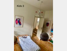 Kinderzimmer mit hellem Holzboden, einem Kinderbett auf der linken Seite und dekorativen Wandbildern. Eine geöffnete Tür führt zu einer kleinen Treppe, und eine schwarze hängende Kugel sowie ein weißer Kronleuchter mit roten Verzierungen sind sichtbar.