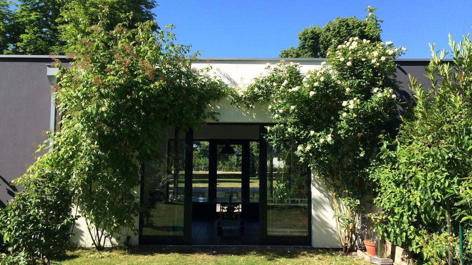 A modern building with dark-framed glass doors and windows, surrounded by lush green plants and flowering shrubs. Behind the building, tall trees and a clear blue sky are visible, with a well-maintained grassy lawn in the foreground.