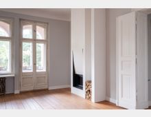 Heller, leerer Wohnraum mit hohen Fenstern und doppelflügeliger Balkontür in weißem Holzrahmen sowie hellem Holzboden. In der Raumecke steht ein moderner, weiß verkleideter Eckkamin mit seitlich gestapeltem Brennholz neben einer hohen weißen Tür.