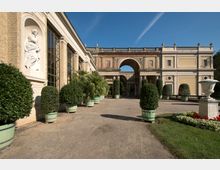 Historische Gartenanlage mit Sandweg, flankiert von symmetrisch angeordneten Zierpflanzen in großen Töpfen. Links ein Gebäude mit großen Fenstern, einer Statue in einer Nische und im Hintergrund ein Torbogen mit klassizistischen Säulen.