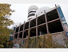 Verfallenes Gebäude mit mehreren offenen Etagen aus Beton und Metallträgern, verziert mit Graffiti. Im Hintergrund sind zwei große, weiße, kuppelförmige Strukturen mit hexagonalem Muster sichtbar, während im Vordergrund Pflanzen und Bäume stehen.