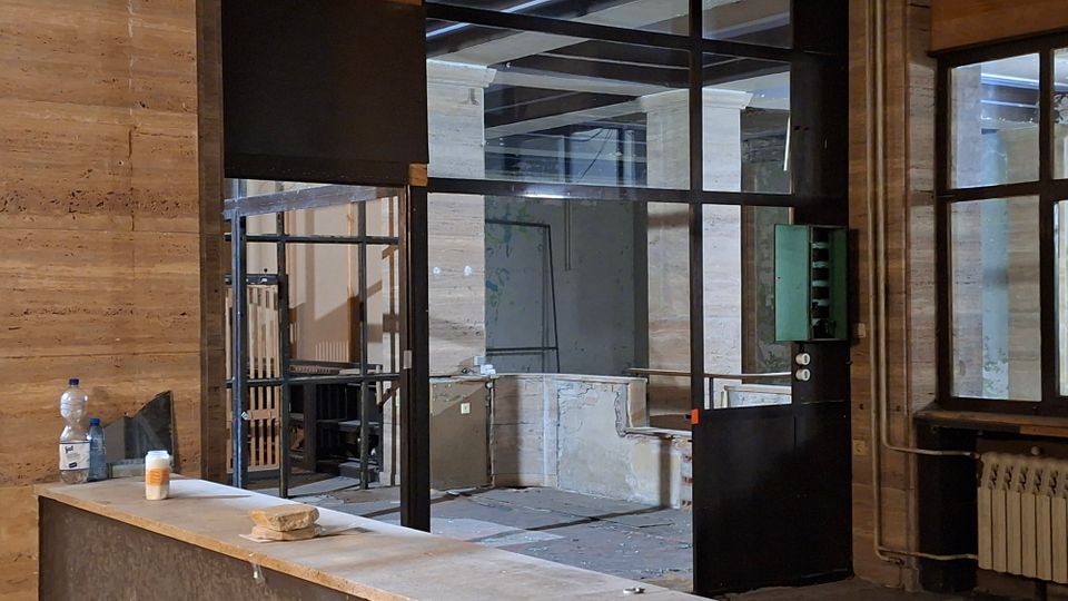 Innenraum eines leerstehenden Gebäudes mit Steinwänden, hohen Säulen und schwarzen Metall-Glas-Trennwänden. Im Vordergrund steht eine staubige Theke mit Flaschen, im Hintergrund sind freiliegende Deckenbalken, abgenutzte Oberflächen und eine Heizung an der Wand zu sehen.