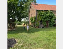 Ein Ziegelgebäude mit rotem Satteldach, teils von grüner Kletterpflanze bedeckt, steht in einer ländlichen Umgebung mit einer grünen Wiese und Bäumen. Im Vordergrund sind unscharf zwei Personen und ein Hund auf der Wiese zu sehen, während eine kleine Holzstruktur links am Rand sichtbar ist.