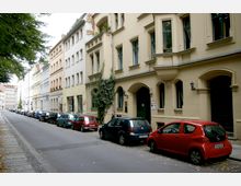 Straßenszene mit historischen Wohngebäuden in beige und hellen Farben, die Fenster mit grünen Rahmen und einige mit Blumenkästen. Auf der rechten Straßenseite sind Autos parallel geparkt, während links ein gepflasterter Gehweg mit Bäumen sichtbar ist.
