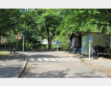 Asphaltierte Fläche einer kleinen Parkanlage mit einem Zebrastreifen, Verkehrsschildern für einen Kreisverkehr und einen Fußgängerüberweg. Im Hintergrund sind Bäume, eine Holz- und eine Metallhütte sowie eine Sitzbank sichtbar.