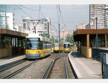 Straßenbahnhaltestelle in einer städtischen Umgebung mit gelb-weißen Straßenbahnen auf parallelen Gleisen. Die Haltestelle ist von überdachten Wartebereichen umgeben, während hohe Wohn- und Bürogebäude im Hintergrund zu sehen sind.