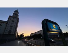 Blick in der Dämmerung auf eine breite Stadtstraße mit mehreren Fahrspuren und angrenzenden Wohn- und Geschäftsbauten. Im Vordergrund steht ein beleuchtetes U-Bahn-Schild „U Frankfurter Tor“, links ragt ein hohes Gebäude mit Turmkuppel in den Himmel.