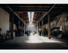 Großer, industrieller Innenraum einer alten Fabrikhalle mit sichtbaren Metallträgern und Holzdecken. In der Mitte führt ein geschmückter Weg zu einer Bühne, während auf beiden Seiten Sitzplätze und Dekorationen mit Pflanzen und Blumen zu sehen sind.