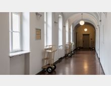 Ein heller Flur mit weiß getünchten Wänden, hohen Fenstern und einer gewölbten Decke. Entlang einer Seite stehen mehrere gelbe Transportwagen mit Drahtgestellen, während die polierten Holzböden und hängenden Kugel-Lampen den Raum definieren.