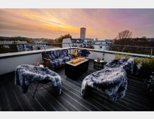 Dachterrasse mit Holzfußboden und einer Sitzecke aus Sesseln und Sofas, die mit grauen Kunstfell-Decken bedeckt sind, rund um einen rechteckigen Gaskamin. Im Hintergrund sind moderne Gebäude, Bäume und ein farbiger Sonnenuntergang sichtbar.