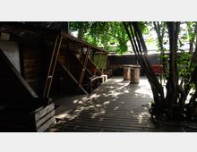 Holzterrasse im Freien mit schattenspendenden Bäumen, Holzstrukturen an der linken Seite und Metallfässern in der Mitte. Der Bereich wirkt wie ein improvisierter Outdoor-Raum in einem urbanen Umfeld.