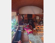 Innenraum eines Wohnraums mit hoher, gewölbter Holzdecke und großen Fenstern auf der linken Seite. Der Raum ist eingerichtet mit einem Essbereich, einer offenen Küche, mehreren Teppichen, Pflanzen und dekorativen Gegenständen auf Regalen und Tischen.