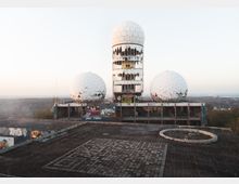 Ehemalige Abhörstation auf dem Teufelsberg in Berlin mit drei großen weißen, kuppelförmigen Radarkuppeln auf einem heruntergekommenen Gebäude, das mit Graffiti verziert ist. Die Umgebung zeigt eine waldreiche Landschaft im Hintergrund.