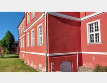 Roter Fassadenabschnitt eines mehrgeschossigen Gebäudes mit weißen Fensterrahmen und Putzstruktur. Im Vordergrund ein kleiner Grünbereich mit Gras, im Hintergrund ein einzelner Baum und blauer Himmel.
