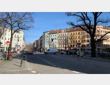 Straßenszene an einer Kreuzung mit Altbauhäusern in unterschiedlichen Pastellfarben, kahlen Bäumen und geparkten Fahrrädern. Im Vordergrund sind eine rote Ampel, ein Verkehrszeichen und ein geparkter Lieferwagen sichtbar.
