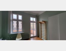 Heller Innenraum mit hohen Fenstern und einer Glastür zum kleinen Balkon, darunter ein Heizkörper und ein Stuhl. Rechts stehen eine Sprossenwand aus Holz und ein großer weißer Schrank vor grün gestrichenen Wänden und Holzboden.
