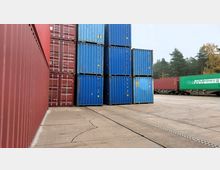 Ein Lagerplatz mit gestapelten roten und blauen Frachtcontainern, daneben Güterwaggons mit weiteren Containern. Im Hintergrund sind Bäume einer Waldlandschaft sichtbar.