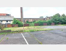 Freifläche mit Asphaltboden und vereinzeltem Grasbewuchs, im Hintergrund ein langgezogenes Backsteingebäude mit vielen kleinen Fenstern, ein Schornstein und dichte Vegetation. Links ist ein Wohnhaus mit roten Dächern sichtbar.