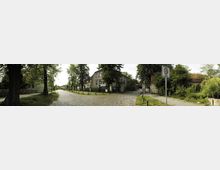 Straßenansicht eines ländlichen Wohngebiets mit Kopfsteinpflaster, flankiert von alten Häusern und Bäumen. Ein Verkehrsschild markiert eine 30-km/h-Zone, und Gehwege aus Schotter verlaufen entlang der Straße.