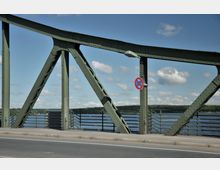 Grüne Stahlbrücke mit Fachwerkstruktur, darunter ein Fluss und im Hintergrund eine Landschaft mit Bäumen. Ein Straßenschild mit Parkverbot hängt an einem der Brückenpfeiler, daneben eine asphaltierte Fahrbahn.