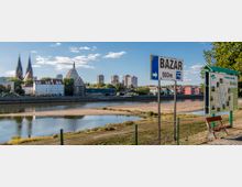 Blick auf die Oder mit Uferbereich im Vordergrund, einer Beschilderung Richtung „Bazar“ und einer Karte von „Frankfurt (Oder) - Słubice“. Im Hintergrund sind Gebäude der beiden Städte sichtbar, darunter markante Kirchtürme und moderne Hochhäuser.