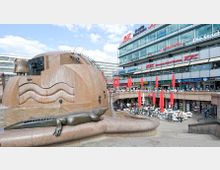 Ein öffentlicher Platz mit einem großen Brunnen im Vordergrund, der eine tierähnliche Skulptur, wellenförmige Elemente und Wasserläufe zeigt. Im Hintergrund befinden sich mehrstöckige Geschäftsgebäude mit Restaurants und Geschäften sowie eine Außenterrasse mit roten Sonnenschirmen und Sitzgelegenheiten.