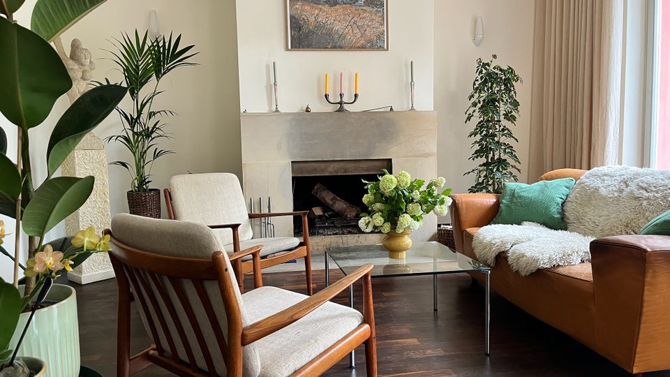 Helles Wohnzimmer mit zentralem, weiß verkleidetem Kamin und einem Landschaftsbild darüber, davor ein Glas-Couchtisch mit Blumenstrauß. Rechts steht ein braunes Ledersofa mit Kissen und Decke neben bodentiefen Fenstern mit Vorhängen; links sind zwei Holzsessel und mehrere große Zimmerpflanzen auf dunklem Holzboden angeordnet.