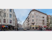 Straßenszene mit mehrstöckigen Wohngebäuden im Eckstil, darunter Geschäfte und Cafés mit bunten Markisen im Erdgeschoss. Im Vordergrund sind parkende Autos, Fußgänger und kahle Bäume zu sehen; der Himmel ist blau mit vereinzelten Wolken.