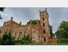 Alte, verfallene Backsteinruine eines historischen Gebäudes mit einem hohen Turm und gotischen Fenstern, teilweise mit Efeu bewachsen, umgeben von Bäumen und Natur.