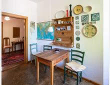 Gemütlicher Essbereich mit kleinem Holztisch und drei Stühlen, einer hängenden Lampe und einer großen Weltkarte an der Wand. Daneben hängen ein rustikales Wandregal mit Keramik sowie dekorative Teller und Fliesen; durch einen Holztürrahmen ist ein angrenzendes Zimmer mit Teppich zu sehen.