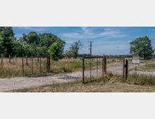 Eine offene Metallzauntür führt auf eine ländliche Wiese mit trockenem Gras und vereinzelten Bäumen. Im Hintergrund sind Strommasten, ein Schild sowie eine Baumreihe am Horizont zu sehen, unter klarem, blauem Himmel.