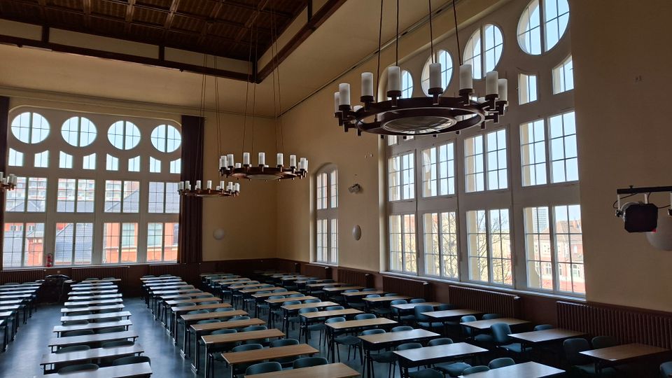 Großer Hörsaal mit hohen Decken, ringförmigen Kronleuchtern und langen Reihen von Tischen und Stühlen. Hohe Fensterfronten mit runden Oberfenstern lassen viel Tageslicht herein und geben den Blick auf umliegende Gebäude frei.