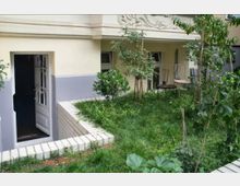 Kleiner grüner Gartenbereich vor einem Gebäude mit hellbeiger Fassade. Eine geöffnete Tür führt in das Gebäude, neben der gepflegten Rasenfläche stehen mehrere junge Bäume und Gartenstühle.